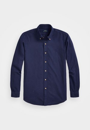 Chemise bleu marine à manches longues avec boutons et col boutonné, boutons beige, posée à plat sur fond blanc.