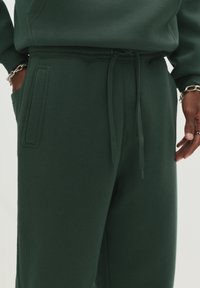 Dunkelgrüne Sweatpants mit Kordelzugverschluss und Seitentaschen, getragen von einer Person mit Armbändern an dunklen Händen.