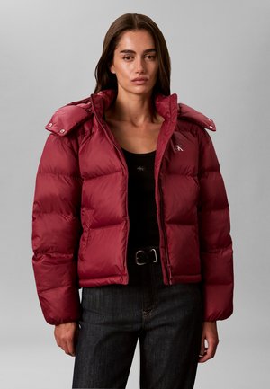 Frau trägt eine rote Daunenjacke über einem schwarzen Top und dunklen Jeans, steht vor einem schlichten grauen Hintergrund.