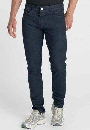 Uomo che indossa jeans slim fit scuri e scarpe da corsa argento-grigie in piedi contro uno sfondo chiaro e uniforme.