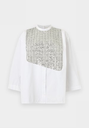 Kvinne iført løs hvit topp med sølvglitrende frontpanel, lys grå midi skjørt med blonderkant og svarte sandaler med hæl, stående.