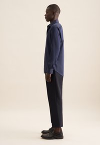 Marineblauwe button-up shirt met lange mouwen, zachte textuur. Gekoppeld aan op maat gemaakte donkere broek en zwarte schoenen, wat een slanke silhouet benadrukt.