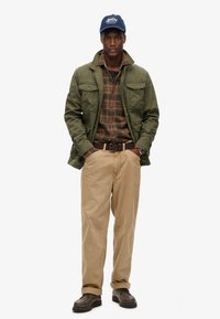Olivengrøn jakke med to brystlommer, ternet skjorte i brune og grønne nuancer, beige bukser, brune sko og en marineblå kasket med logo.