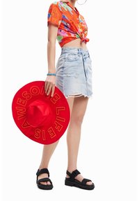 Chapeau à large bord rouge avec "LA VIE EST INCROYABLE" en lettres jaunes, chemise courte à fleurs orange, jupe en denim déchirée, et sandales noires.