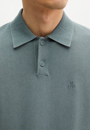 Uomo che indossa una polo azzurra chiusa con bottoni, in tessuto strutturato, con un piccolo logo ricamato sul petto, ritratto dall'alto del collo in giù.