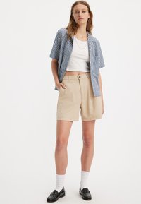 Shorts en coton beige avec une coupe décontractée, dotés de poches avant et d'une fermeture à boutons. Portés avec une chemise à manches courtes à carreaux bleu et blanc.