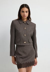 Vrouw in een bruine, gebreide cropped jack met grote gouden knopen en bijpassende rok met kanten zoom, staand tegen een effen witte achtergrond.