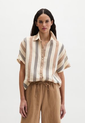 Frau mit langen dunklen Haaren, die ein beige kurzärmliges Hemd mit vertikalen braunen und schwarzen Streifen sowie lockere braune Hosen mit Kordelzug trägt.