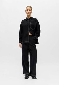 Femme debout portant un pantalon noir ample et une chemise noire oversize assortie avec des coutures blanches, une main dans la poche, fond neutre.