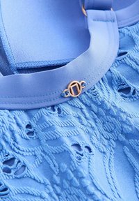 Hellblauer Stoff mit aufwendiger Stickerei und ausgeschnittenen Mustern. Der Ausschnitt hat ein glattes Band mit einem goldenen, runden Verschlussdetail.