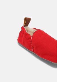 Czerwony materiałowy pantofel z miękkim, kremowym wyściółką, z brązową skórzaną pętlą przy pięcie i płaską, szaro-outsole.