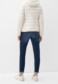 Lys beige puffjakke med hætte, præget af quiltet tekstur. Sammen med mørkeblå jeans og hvide sneakers. Simpelt design, afslappet look.