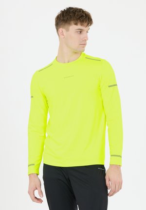 Man in een gele lange mouwen sportshirt met reflecterende strepen en zwarte broek kijkt naar rechts tegen een witte achtergrond.