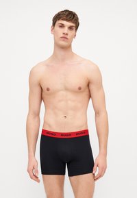 Boxer-briefs noirs avec une taille élastique rouge arborant la marque "HUGO". Le matériau semble lisse ; le design ajusté met en valeur la silhouette.
