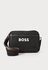 Čierna crossbody taška vyrobená z textúrovanej látky, obsahujúca prednú zipsovú kapsu, nastaviteľný popruh a výrazné biele logo vpredu.