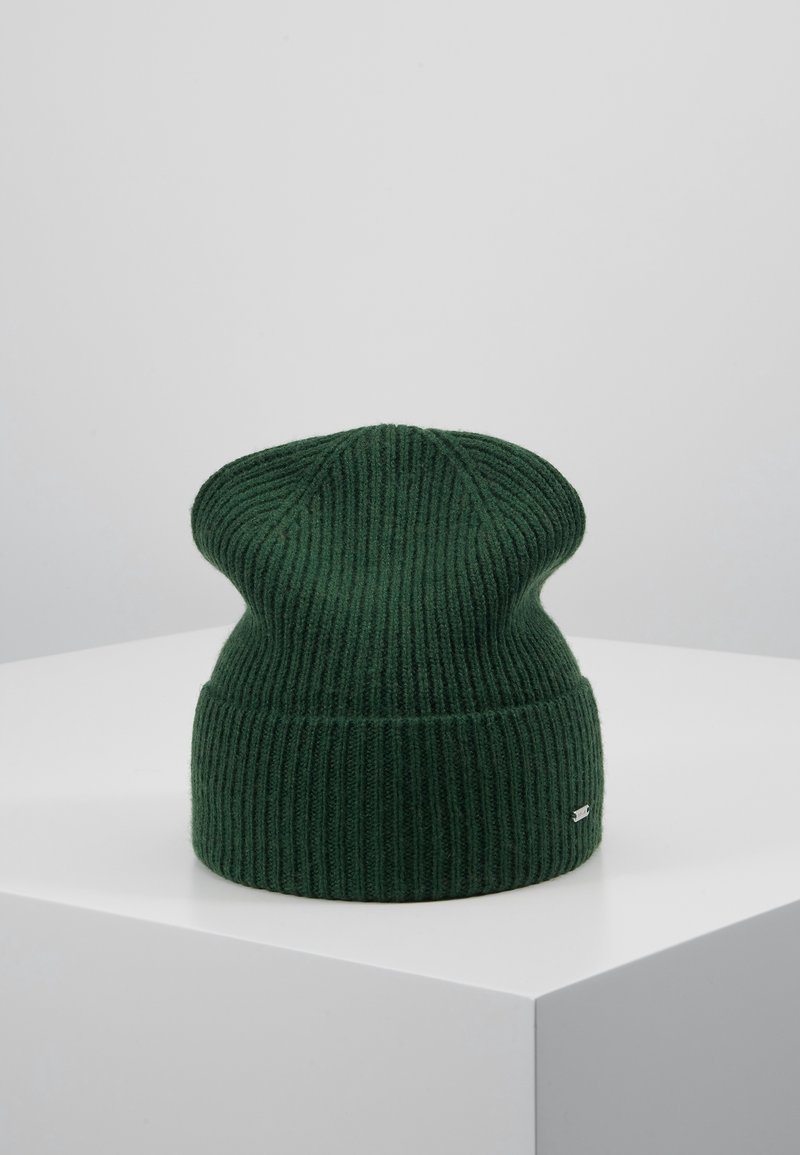 Bonnet en tricot côtelé vert foncé avec un revers plié, exposé sur un socle blanc sur fond gris clair.