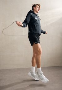 Czarny hoodie z szarymi akcentami, czarne szorty, białe skarpetki i szaro-białe buty sportowe. Osoba skacząca z liną w trakcie skoku w pomieszczeniu.