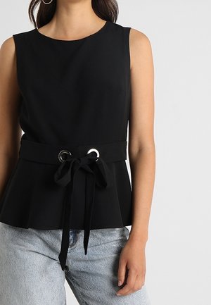 Czarny top bez rękawów z okrągłym dekoltem, talią z baskinką oraz materiałowym paskiem przewleczonym przez srebrne oczka, noszony z jasnoniebieskimi dżinsami.