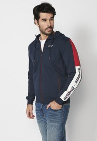 Jūrinės mėlynos zip-up džemperis su raudonais pečių akcentais ir baltais rankovių užrašais. Įrengtas su raišteliu reguliuojamu kapisonu ir dviem šoninėmis kišenėmis. Tekstūruota medžiaga.