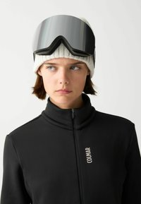 Crna zip-up jakna s visokim ovratnikom, izrađena od teksturirane tkanine. Model nosi bijelu pletenu kapu i velike zrcalne skijaške naočale.