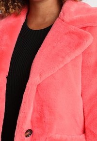 Pink faux fur jacket with a wide lapel, single button closure, and soft texture, paired with a ribbed black sweater underneath.