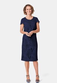 Navyblauwe jurk met korte mouwen, bloemen uitsnijdingen, knielengte, gemaakt van textuurstof. Model staat lachend, draagt bijpassende hakken.