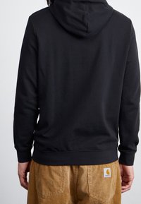 Persona che indossa un felpa nera con cappuccio e pantaloni di velluto a coste marroni con il logo Carhartt applicato sulla tasca, mostrata di spalle su uno sfondo semplice.