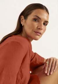 Roestkleurige linnen top met losse mouwen. Draagt gouden oorbellen en een gelaagde ring. Glad haar en neutrale nagellak.