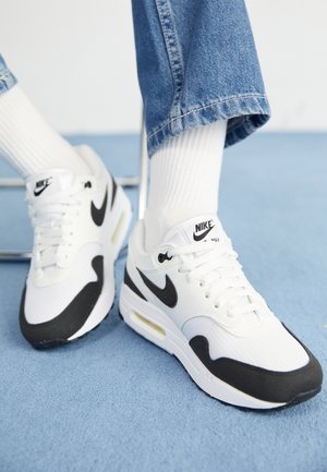 Osoba nosząca białe i czarne sneakersy Nike Air Max z białymi skarpetkami i niebieskimi dżinsami, stojąca na niebieskim wykładzinowym podłodze.