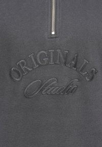 Dunkelgrau sweatshirt mit Reißverschluss, das geprägte Texte mit der Aufschrift "ORIGINALS" und "Studio" in kontrastierendem Faden zeigt. Glatte Textur mit metallischem Reißverschluss.