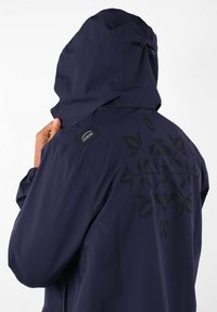 Veste à capuche navy, arborant un motif en relief de feuilles noires dans le dos, avec une étiquette de logo Oxbow sur l'épaule. Tissu lisse et résistant à l'eau.