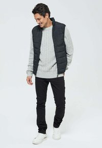 Gilet matelassé noir sur un pull en maille torsadée gris clair, associé à un jean slim noir et des baskets blanches ; tenue décontractée avec des textures superposées.