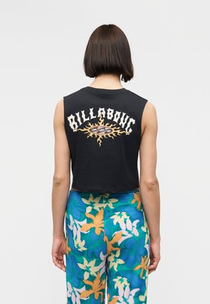 Persona con capelli castani corti che indossa un top nero senza maniche con un logo sole e la scritta "Billabong", e pantaloni con un vivace motivo floreale.