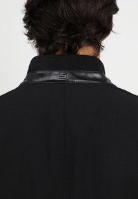 Giacca nera con tessuto testurizzato, colletto in pelle e un piccolo dettaglio del logo nella parte posteriore del collo.