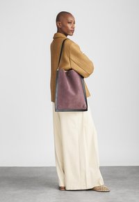 Liela somu soma tumši violetā zamšā ar melnu ādas apdari. Kvadrātveida forma ar minimālistisku dizainu. Nēsāta uz pleca ar teksturētu džemperi.