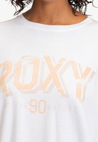 Bílý bavlněný tričko s velkým, vybledlým grafickým potiskem ve světle oranžové barvě a textem "ROXY" a "90 ACTIVE NATURE" v tučném designu.