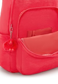 Světlý růžový batoh vyrobený z lehké tkaniny, s přední kapsou, jasným zipem a připevněným huňatým červeným klíčenkou opičky.