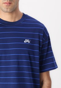 Tamsiai mėlynos spalvos medvilninė marškinėlių su horizontaliomis šviesiai mėlynomis juostelėmis, trumpomis rankovėmis ir mažu baltu Nike SB logotipu ant krūtinės.