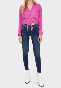 Camicia rosa con bottoni e maniche lunghe, legata in vita. Jeans blu scuri con leggera abrasione, abbinati a stivaletti bianchi alla caviglia.