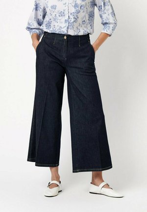 Frau trägt dunkle, weit geschnittene, kurz geschnittene Jeans, weißes Hemd mit Blumenmuster und weiße Mary-Jane-Ballerinas mit Schnallen. Hände in den Taschen.