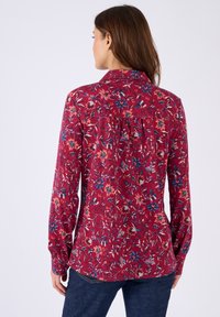 Donna vista di spalle che indossa una camicia rossa a maniche lunghe con un motivo floreale multicolore e jeans blu scuro, su uno sfondo semplice.