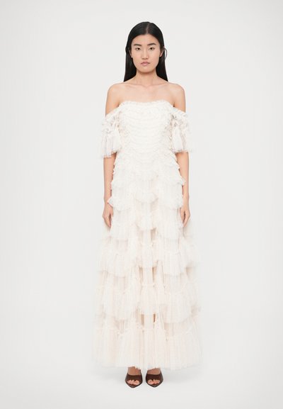 Robe blanche à épaules dénudées avec des volants superposés, comprenant des détails en tissu transparent et un corsage ajusté, associée à des escarpins ouverts foncés.