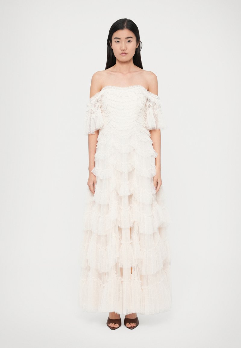 Off-the-shoulder white dress with layered ruffles, featuring sheer fabric details and a fitted bodice, paired with dark open-toe heels.