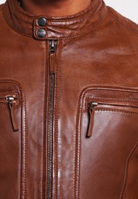 Chaqueta de cuero marrón con un alto cuello, cierre central y dos bolsillos con cremallera. Textura suave con detalles de costura sutiles.
