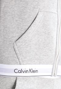 Hanorac gri cu buzunar frontal, având un bandou cu logo alb Calvin Klein și închidere cu fermoar pe lateral. Textură moale.