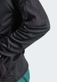 Svart atletisk jacka med ett texturerat tyg och en ficka med dragkedja på överarmen. Detaljer inkluderar kontrasterande mörka accenter på ärmen.