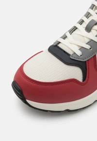 Red and white sneaker with a mesh body, rubber sole, and gray accents. Features flat laces, a textured surface, and rounded toe design.