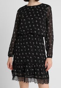 Černé květinové šaty s dlouhými průhlednými rukávy, kulatým výstřihem, nabraným pasem a volánkovým lemem s krajkovým zakončením. Obsahuje malé vícebarevné vzory.