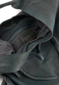Zielona skórzana torba o miękkiej fakturze, z wewnętrzną kieszonką na zamek i metką z napisem "Skórzany produkt wysokiej jakości". Strukturalny design z wygiętymi akcentami.