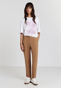 T-shirt bianca in versione oversize con stampa grafica rosa, abbinata a pantaloni tapered beige e sneakers bianche con suola marrone.